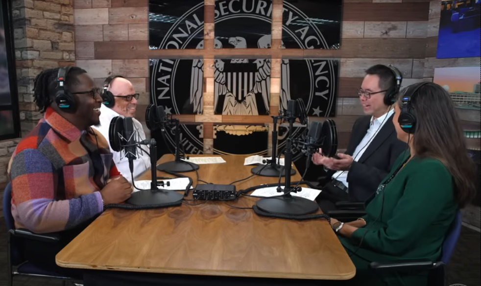 NSA, No such Podcast
Vier Personen sitzen sich an einem Tisch gegenüber. Sie haben alle Podcast Mikrofone vor sich und Kopfhörer auf. Im Hintergrund ist das Logo der NSA zu sehen